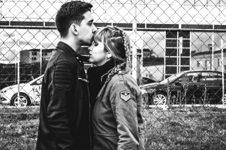 grayscale photo of man kissing forehead of woman near fence photo