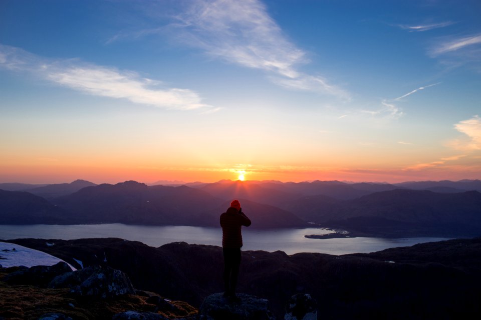 person standing on mountain ridge over lake under cloudy sky at sunset