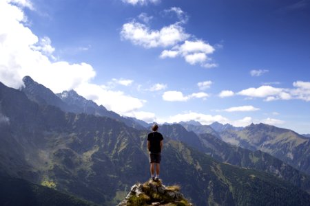 man wearing black shirt and gray shorts on mountain hill beside mountains under white and blue cloudy skies photo