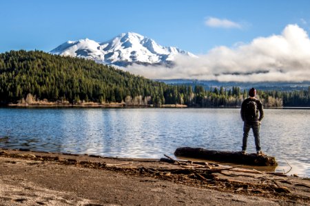 man standing beside body of water near green high trees and white mountain under blue and white sky during daytime photo