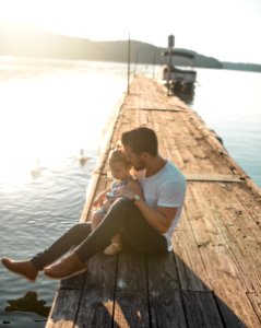 man and girl sitting on brown dock near boat and two white ducks during daytime