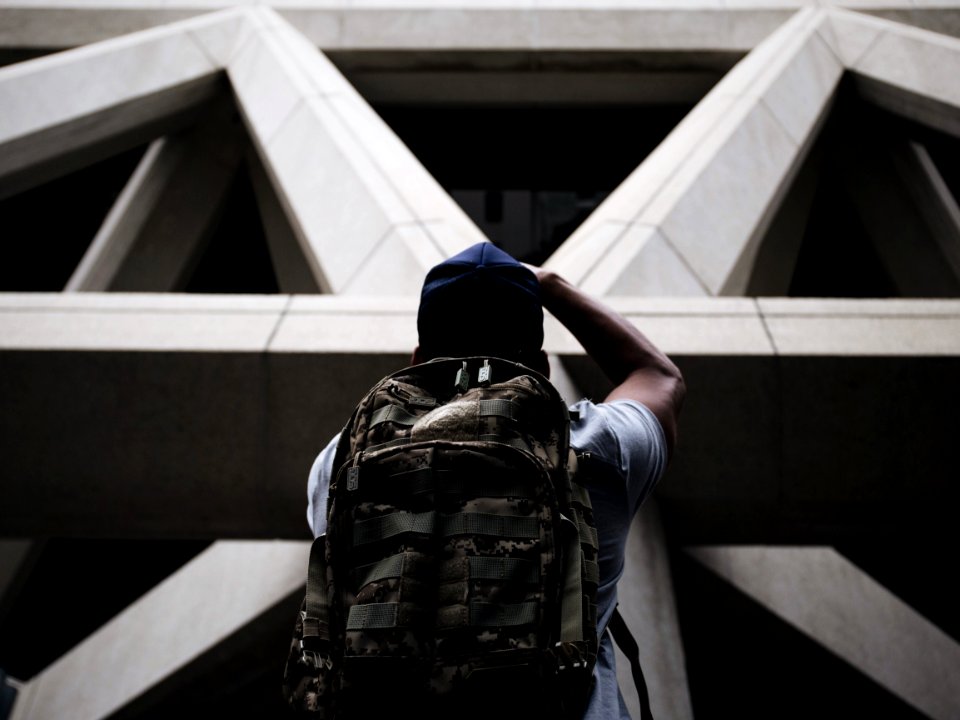 person wearing shirt and hiking bag looking in front of grey concrete structure while standing during daytime