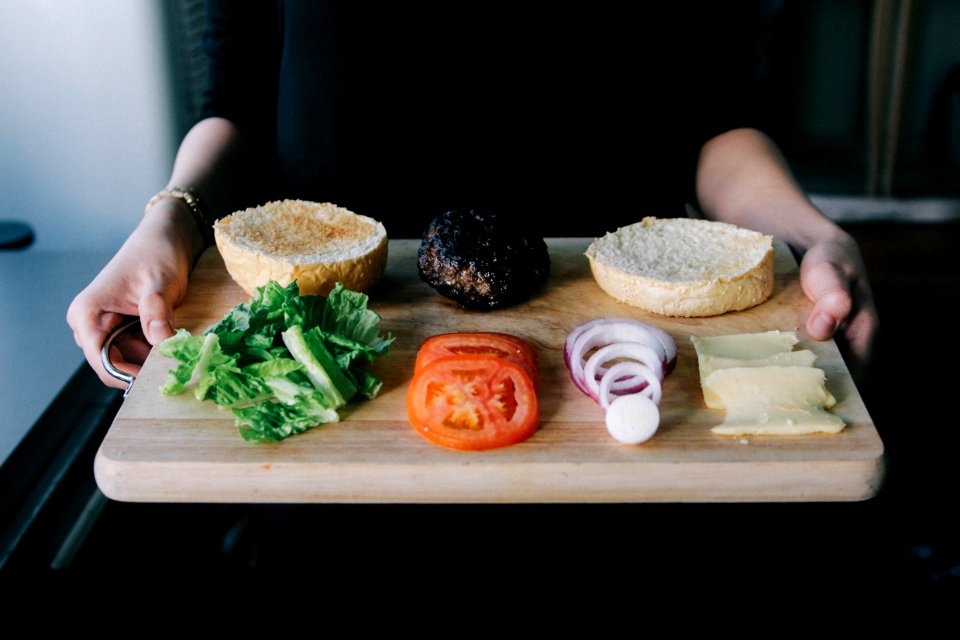 woman holding up wooden tray with lettuce, tomato, onions rings and cooked patty for burger