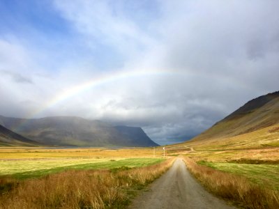 dirt path between brown and green field leading to a rainbow under blue and white cloudy sky photo
