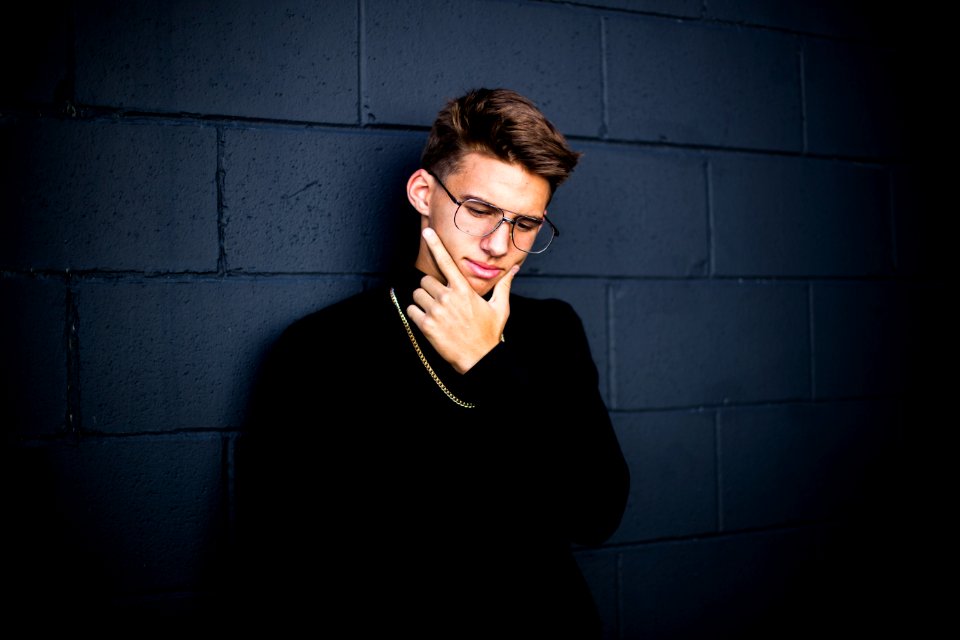 man leaning on wall while looking down wearing eyeglasses and necklace with right hand on chin
