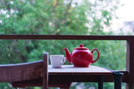 selective focus photo of red ceramic teapot and white ceramic teacup on brown wooden table near tree during daytime photo