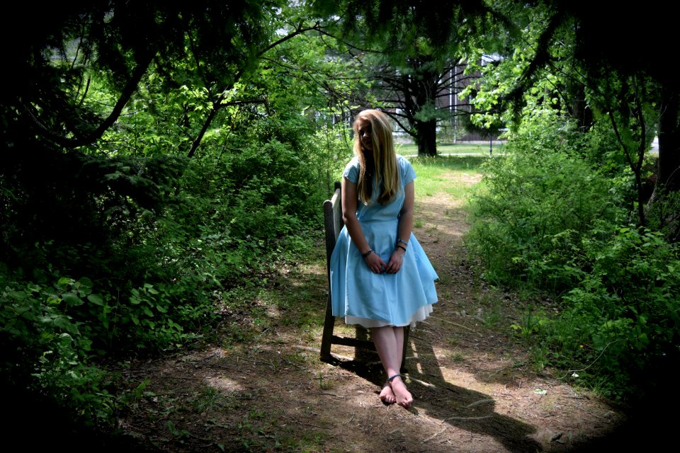 A young adult female in a white dress leaning on a chair in the middle of the woods.