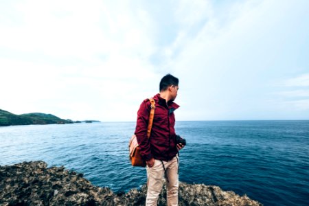 man wearing red zip-up jacket standing on top of rocky cliff beside body of water during daytime photo