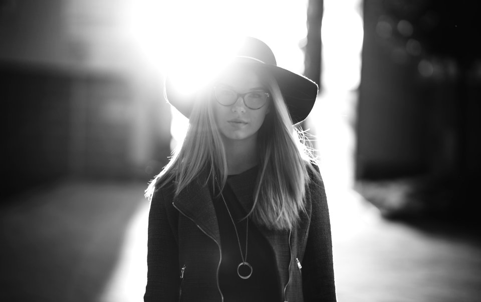 grayscale photography of woman wearing hat and coat standing near wall