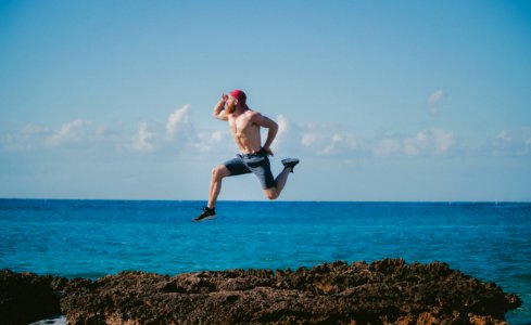 man jumping in mid air while looking ahead beside body of water during daytime photo