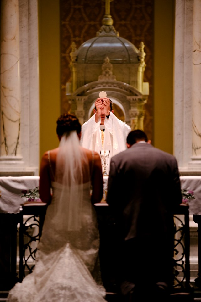 groom and bride kneeling in front of priest raising The Holy Sacrament
