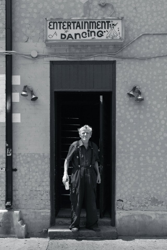 grayscale photo of man wearing collared tops and dress pants standing between concrete wall near Entertainment Dancing signage building photo