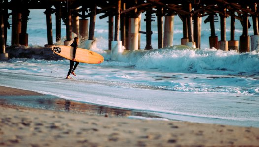 person holding surfboard standing on seashore in front of wave near wooden dock photo
