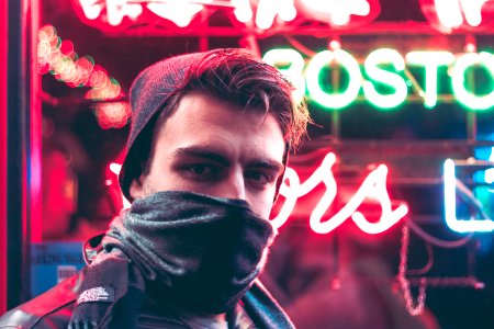 man with face half covered with mask standing in front of red and green neon light signage during daytime