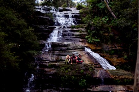 three person sitting on rock formation in waterfalls surrounded with trees at daytime photo