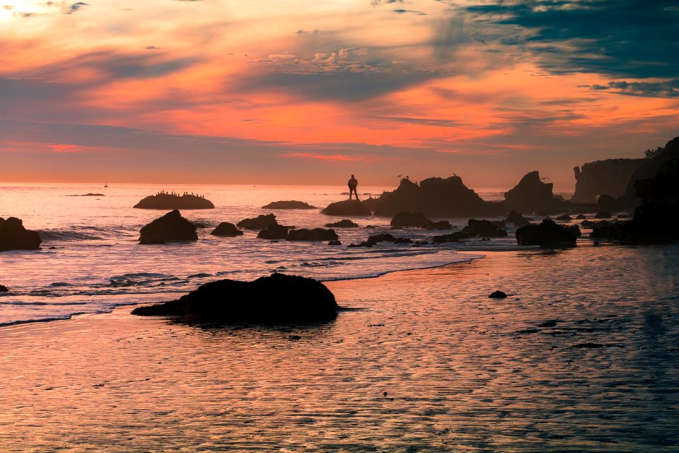 silhouette person standing on seashore rock during daytime