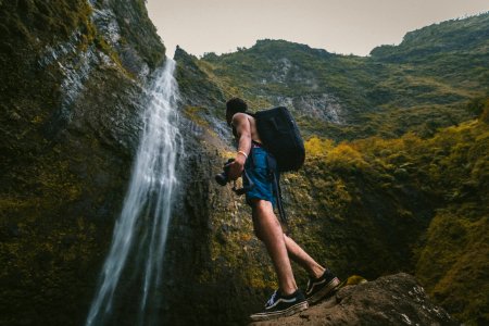 man wearing blue shorts standing on rock formation while holding DSLR camera in front of waterfalls photo