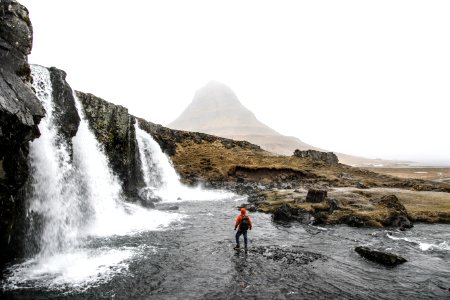 man with backpack standing on river beside waterfalls