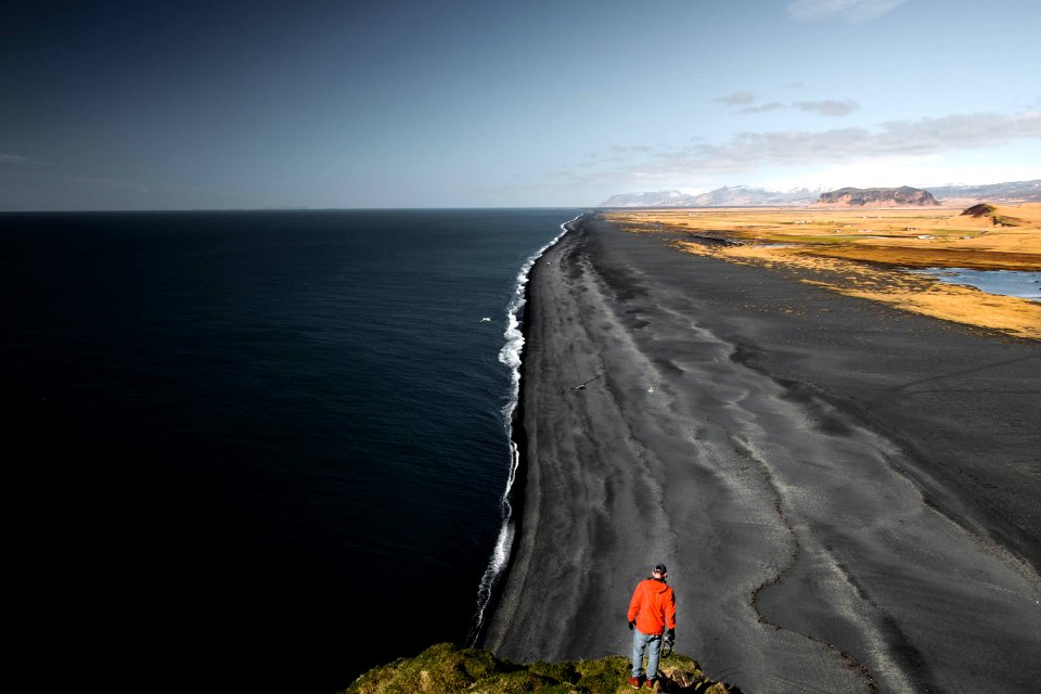 man in red top standing near cliff overlooking seashore during daytime photo