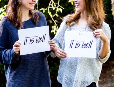 Two women holding up pieces of paper that say "It is well," while looking at each other and smiling. photo