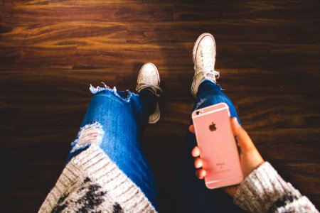 person holding rose gold iPhone 6s wearing distressed blue denim jeans standing on brown wooden floor photo