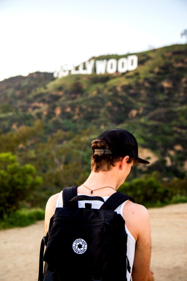 man wearing white tank top and black snapback cap carrying black backpack in distance hollywood signboard at daytime