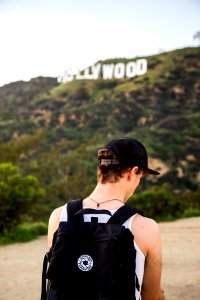 man wearing white tank top and black snapback cap carrying black backpack in distance hollywood signboard at daytime photo