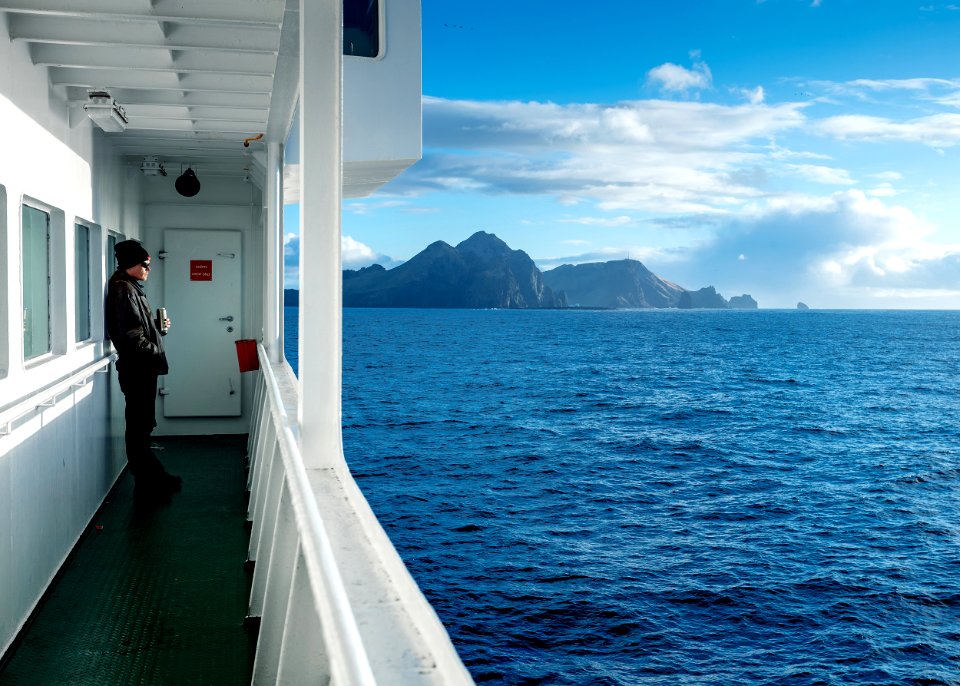 human in gray long-sleeved top wearing black knit cap leaning on boat window while looking on ocean water during daytime