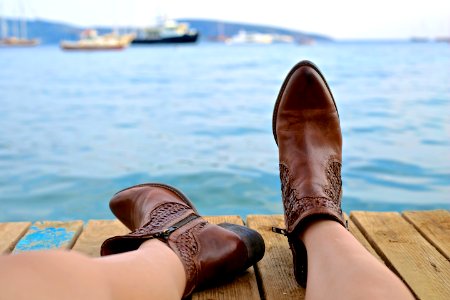 person wearing brown leather side-zip boots sits on brown wooden pier near body of water during daytime photo