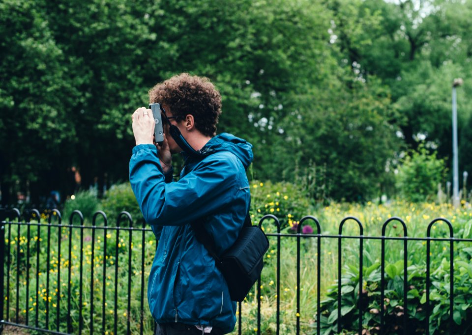 man wearing blue jacket standing and using camera beside flower field surrounded with tall and green trees