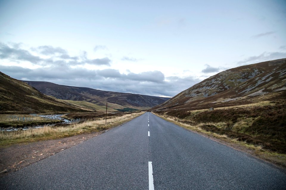 gray concrete road between green grass field near mountain under white clouds during daytime photo