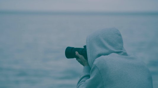 person wearing hoodie holding camera taking photo near body of water during daytime photo