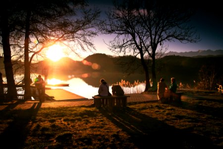 People Sitting on Bench Near Lake and Bare Trees Under Sky during Golden Hour photo