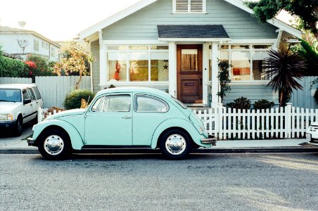 This is a photo of a light mint green old VW beetle car in front of a wodden house with a white fence in a residential area in a typical american street. There is a yucca palm tree in the lawn of the house. The sun is shining brightly. photo