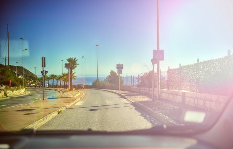 This picture is taken from inside a car through the windhield with the dashboard visible on the lower edge of the picture. The car is driving down a palm-lined ocean road under a blue sky with the ocean inthe background. The sunshine is reflected in the windshield of the car. photo
