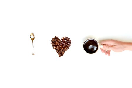 Person Holding Cup Filled With Coffee Beside Coffee Beans Forming Heart and Spoon photo