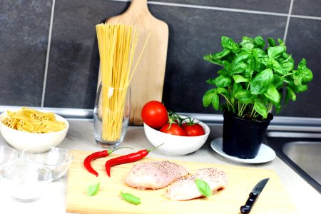 Raw Chicken Breast Seasoned With Peppers Beside Red Chili, Basil, Bowl of Tomatoes, and Raw Pastas on Table photo