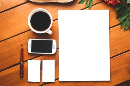 Top view composition of modern smartphone placed near cup of aromatic americano and surrounded with stationeries including blank A4 format paper pen and business card size papers on wooden table