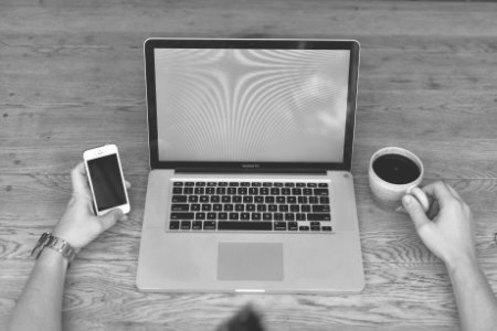 Person Holding Iphone 5 and Cup of Coffee in Front of Macbook Pro Grayscale Photography photo