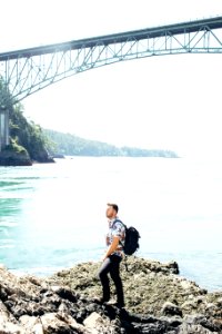 Man Standing on Gray Rock Near Gray Bridge Above Body of Water