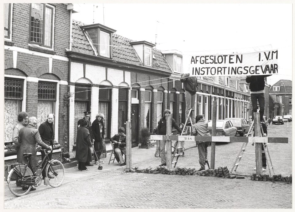 Bewoners in de Romolenstraat brengen een wegversperring aan om het zware verkeer te weren en zo te voorkomen dat de toestand van de verzakte huizen nog verder verslechtert. NL-HlmNHA 54014856