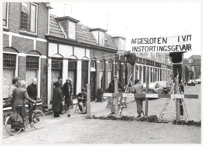 Bewoners in de Romolenstraat brengen een wegversperring aan om het zware verkeer te weren en zo te voorkomen dat de toestand van de verzakte huizen nog verder verslechtert. NL-HlmNHA 54014856