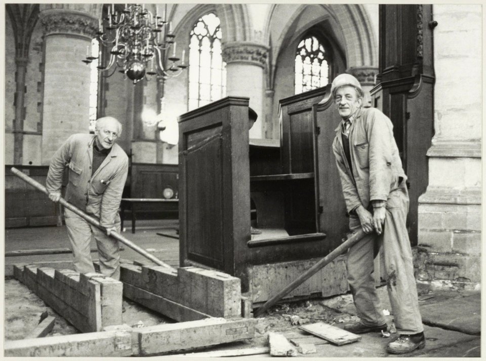 De meubilair restaurateurs Gerard Verbij (links) en Hans van Schaik krikken een kolombank op in de Grote of St. Bavokerk. NL-HlmNHA 54012339