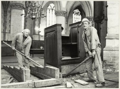 De meubilair restaurateurs Gerard Verbij (links) en Hans van Schaik krikken een kolombank op in de Grote of St. Bavokerk. NL-HlmNHA 54012339