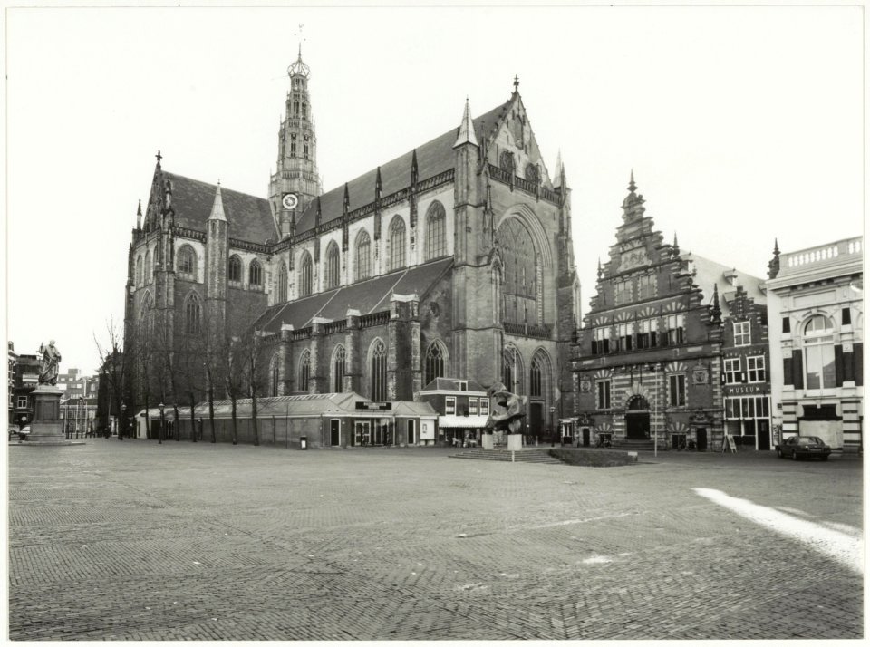 De Grote of St. Bavokerk, rechts van de kerk de Vleeshal. Voor de kerk het beeld De Zonnevechter. NL-HlmNHA 54037586