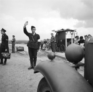 Een man in uniform met armband steekt zijn hand omhoog Hij staat bij een halte, Bestanddeelnr 900-6222