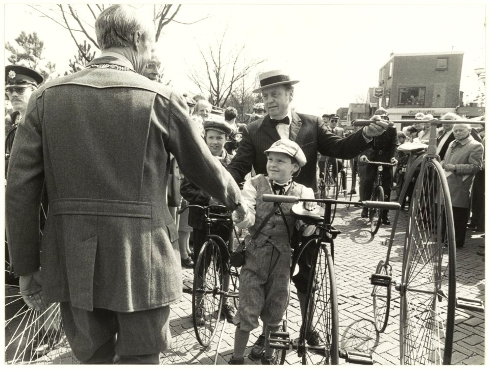 Een moment uit de historische wieleroptocht te Bennebroek. Burgemeester J. de Widt (op de rug gezien) geeft op de Bennebroekerlaan een jonge deelnemer met velocipède de hand. Oriëntatie zie, NL-HlmNHA 54016679