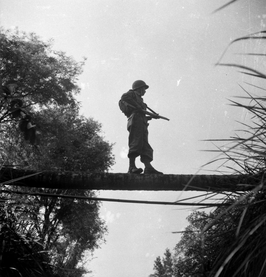 Een militari taat op een boomstam over een beek of sloot, met zijn wapen gerich, Bestanddeelnr 15814