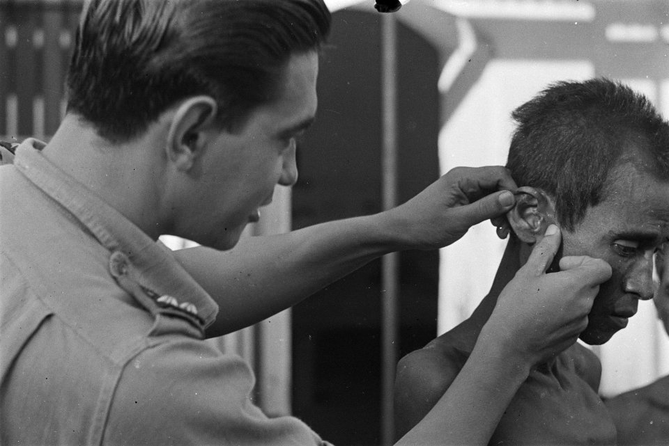Een Nederlandse militaire arts controleert de oren van een Indonesische man, Bestanddeelnr 125A-4-6