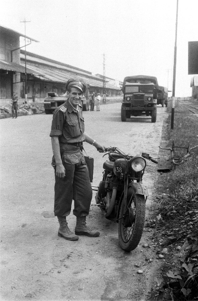 Een sergeant lacht naar de fotograaf, achter hem loodsen en vrachtwagen (Tandjon, Bestanddeelnr 5970